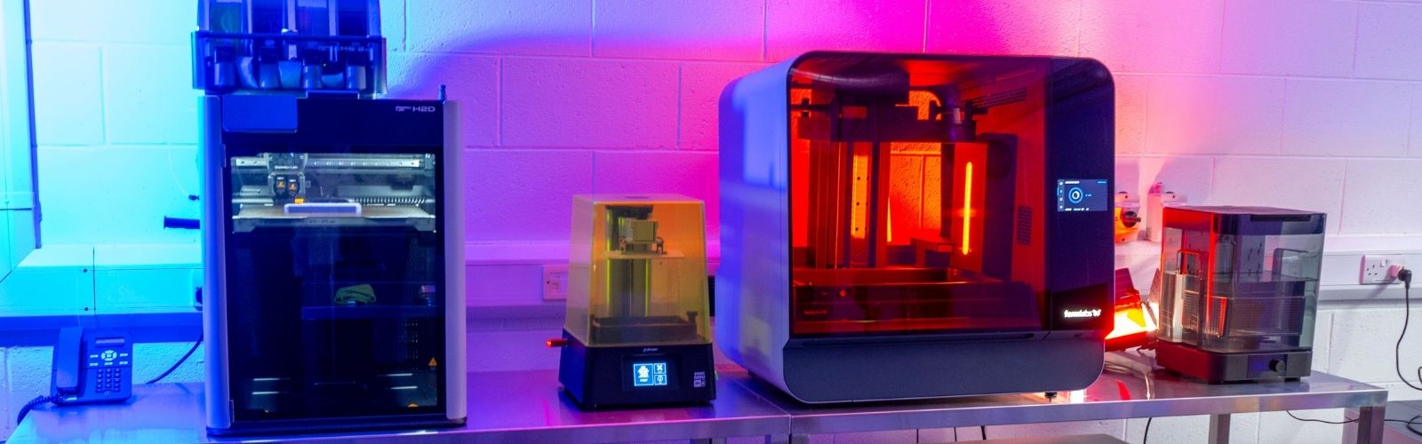 An image of three 3D printers on a bench with red lights in the background