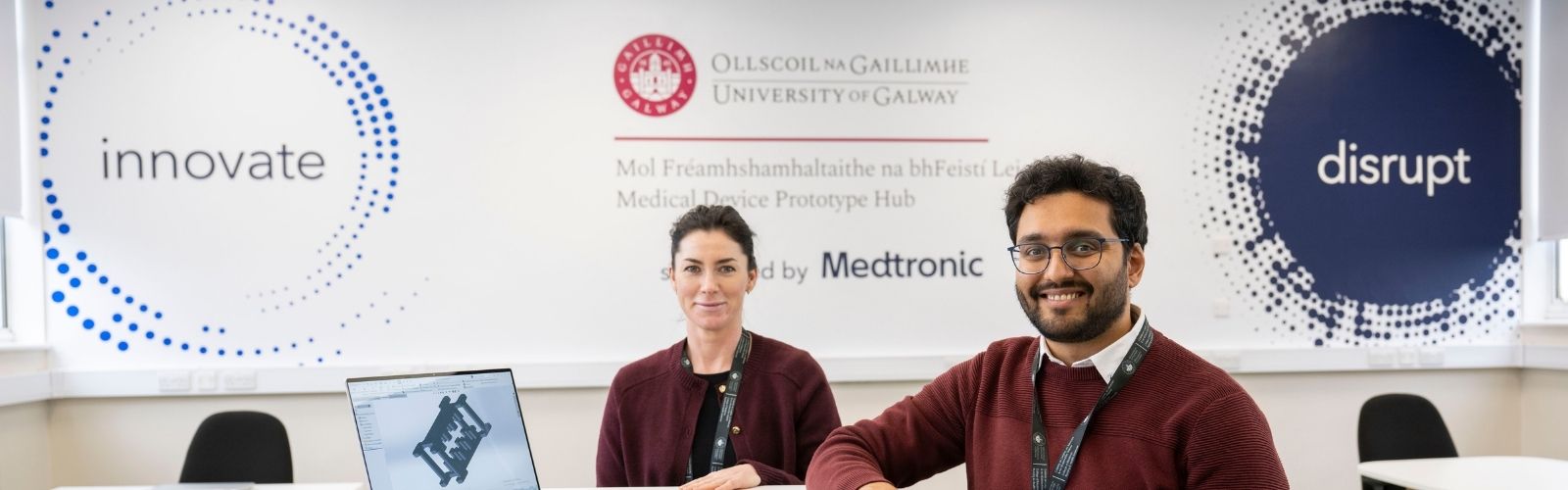 Two people sitting at desks in front of a wall with logos of the University and Medtronic
